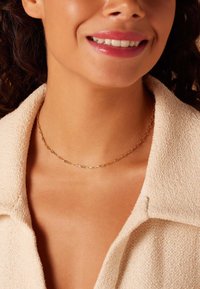 Gouden ketting met langwerpige schakels. Draagt dicht bij de nek, tegen een lichtbeige gestructureerde jas. Gladde metallic afwerking, eenvoudig ontwerp.