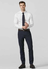 Wit overhemd met kraag, marineblauwe pantalon en een marineblauwe stropdas met stippen. Een leren riem in bruin en schoenen met een donkere print maken de outfit compleet.