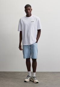 Mann steht vor einer schlichten Wand, trägt ein weißes T-Shirt, hellblaue Jeansshorts, weiße Socken und beige Sneaker, eine Hand in der Tasche.
