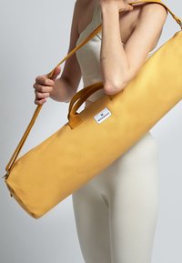 Bolso amarillo alargado con dobles asas y una correa ajustable. Hecho de tela con una textura suave. Presenta una pequeña etiqueta con el logo.