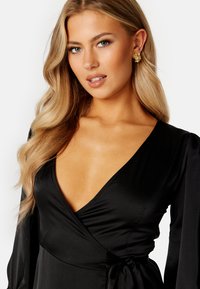 Černé zavinovací šaty s dlouhými rukávy a hlubokým výstřihem do V. Hladký, lesklý materiál s pásem v oblasti pasu. Modelka nosí zlaté květinové náušnice.