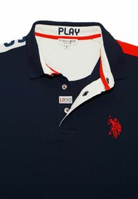 Navy polo shirt with red accents. Features a white collar, embroidered logo, and two-button placket. Made from woven cotton blend.