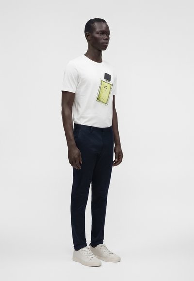 Homme portant un t-shirt blanc avec un grand graphisme jaune de flacon de parfum, pantalon sombre et baskets blanches, debout devant un fond uni.