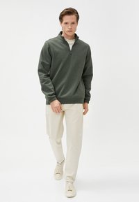 Joven caminando hacia adelante con un suéter de cremallera corta verde oscuro, pantalones crema y zapatillas blancas frente a un fondo blanco plano.