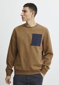 Sudadera de felpa marrón con un bolsillo azul marino, cuello redondo, puños y dobladillo acanalados, y tela texturizada. Diseño casual con detalles en las costuras laterales.