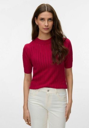 Ženska z dolgimi rjavimi lasmi, oblečena v rdeč pleten top z kratkimi rokavi in bele hlače z visokim pasom, stoji pred enobarvnim svetlim ozadjem.