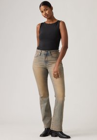 Γυναίκα που στέκεται μπροστά σε απλό φόντο, φορώντας μαύρο τοπ χωρίς μανίκια, ξεθωριασμένο μπλε τζιν και μαύρα μποτάκια αστραγάλου.