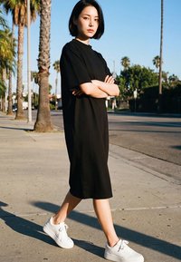 Ung kvinde i en sort kjole og hvide sneakers, der går på en solrig fortov med palmer og bymæssige vejskilte.