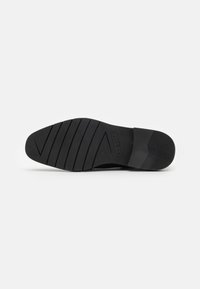 Suela de zapato de goma negra con un patrón texturizado, con ranuras horizontales y verticales para mejorar el agarre y un logotipo grabado en el lateral.