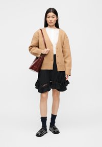 Béžový oversize kardigán s gombíkmi, biela plisovaná blúzka, čierna sukňa s volánikmi a čierne mokasíny; doplnené o bordovú kabelku na rameno.