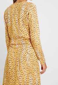 Kvinna iklädd en långärmad klänning i gult och brunt med prickar, sedd bakifrån mot en enfärgad ljusgrå bakgrund.