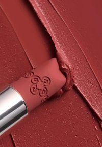 Matowa pomadka w delikatnym różowym odcieniu, o kształcie kuli, z wygrawerowanym logo. Kremowa tekstura pokazuje rozmazaną aplikację na powierzchni.