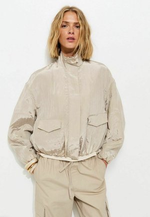 Mujer con cabello rubio a la altura de los hombros lleva una chaqueta bomber beige brillante y pantalones beige claro, de pie con las manos en los bolsillos contra un fondo blanco.