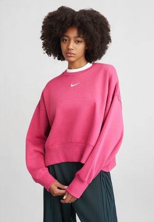 Rozā pieguļošs džemperis ar garām piedurknēm un ribotām malām. Uz krūtīm ir balts Nike logotips. Apvienots ar tumšām svārku biksēm.