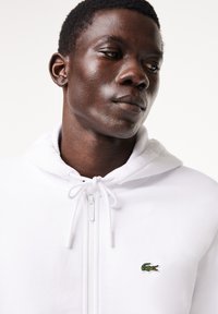 Vit zip-up hoodie med dragsko och huva, med en liten grön krokodil-logotyp på vänster bröst. Mjukt tyg och enkel design.