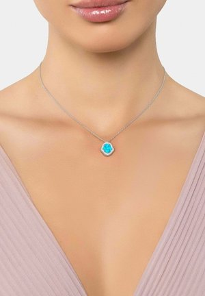 Collier chaîne en argent avec un pendentif turquoise en forme de goutte entouré de petites pierres claires, porté avec un haut rose pâle à col en V.