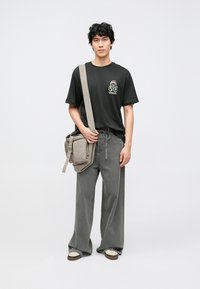 Musta t-paita, jossa vihreä graafinen logo, harmaat leveälahkeiset housut ja harmaa olkalaukku. Beigejä tennareita ruskeilla pohjilla. Yksinkertainen, rento malli.