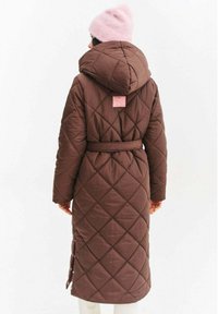 Osoba stojaca chrbtom, oblečená v dlhej prešívanej hnedej bunde s kapucňou a opaskom, tiež má na hlave svetloružovú pletenú čiapku.