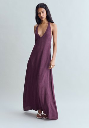 Femme portant une robe violette sans manches, longue jusqu'au sol, avec un décolleté en V profond, et des sandales blanches, debout devant un fond gris clair uni.