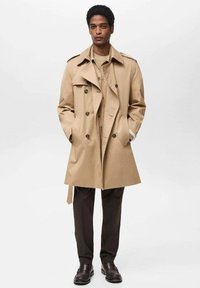 Béžový trench coat s dvojakými gombíkmi, širokými klopami a ramennými epauletami. Nosený cez hnedú košeľu a tmavé nohavice, stojí.