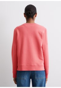 Korallilillane sweatshirt, millel on ümar kaelus, pikad varrukad ja soonikkoes mansetid. Silmade tekstuur ja lõdvestunud istuvus, koos siniste teksadega.