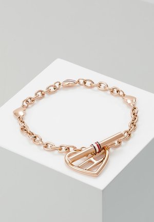 Bransoletka z łańcuszkiem w kolorze różowego złota z zapięciem w kształcie serca i małymi ogniwami w kształcie serduszek, prezentowana na białym podium na szarym tle.