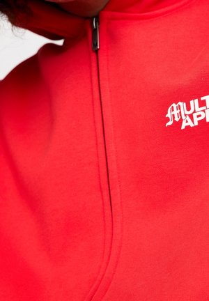 Zbliżenie czerwonej bluzy z zamkiem i częściowym białym napisem "MULTI APP" na piersi, skupiające się na zamku i fakturze materiału.