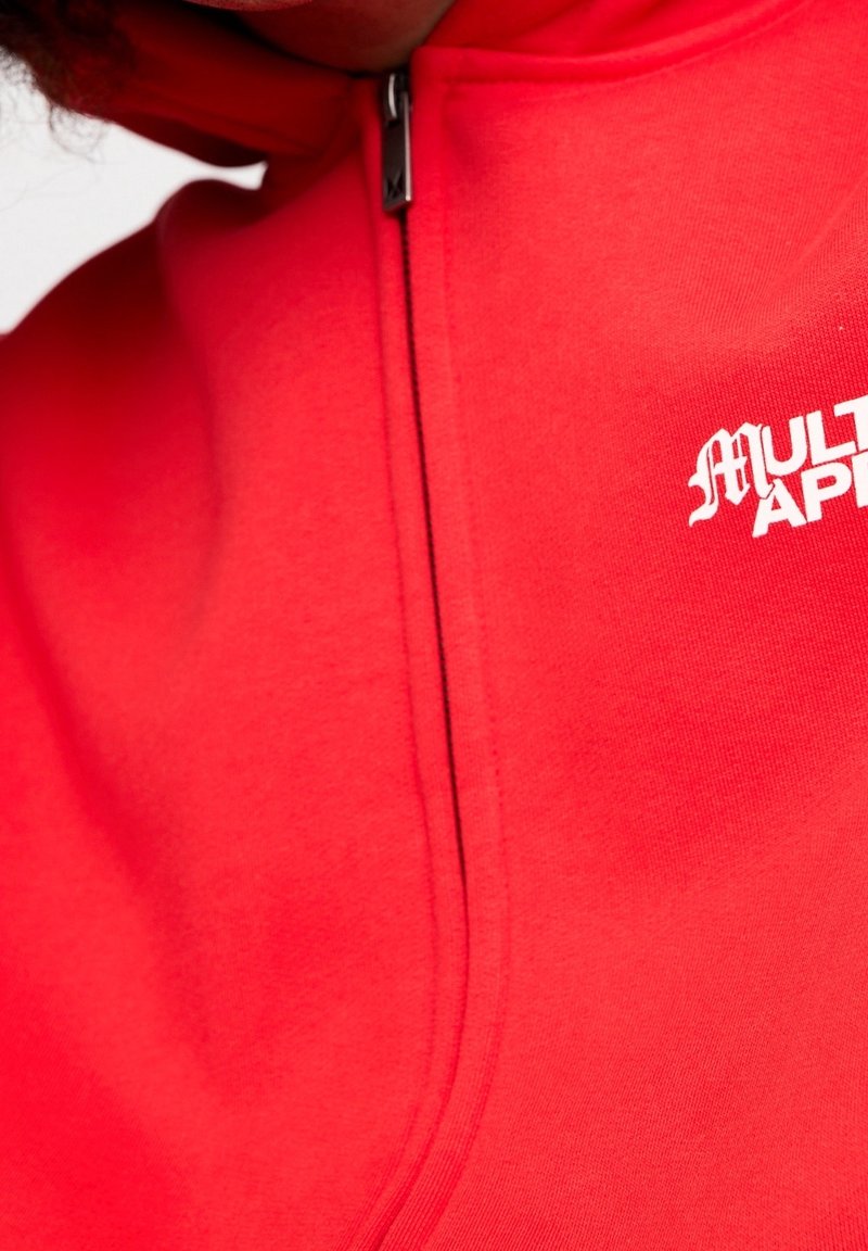 Gros plan sur un sweat à capuche rouge zippé avec un texte blanc partiel "MULTI APP" sur la poitrine, mettant en valeur la fermeture éclair et la texture du tissu.