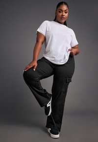 Vit t-shirt med spetsdetaljer vid ärmarna, kombinerad med svarta cargobyxor med sidofickor. Modell i mörka sneakers. En enfärgad grå bakgrund.