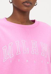 Rosa Sweatshirt mit geripptem Halsausschnitt, mit dem Text „MILANO“ in weißen fetten Buchstaben und „ITALIA“ darunter. Weiche Stoffstruktur.