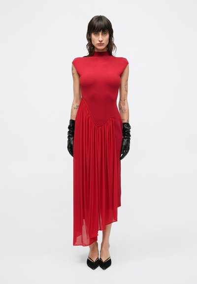 Rotes Kleid mit Kappenärmeln, tailliertem Oberteil und fließendem asymmetrischem Saum. Der Stoff kombiniert feste und durchsichtige Texturen. Schwarze Handschuhe und spitze Schuhe.
