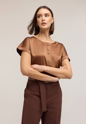 Frau mit kurzen braunen Haaren trägt eine glänzende bronzefarbene Bluse, hoch taillierte braune Hose mit Gürtel, goldene Creolen und lange goldene Halskette, die Arme verschränkt.