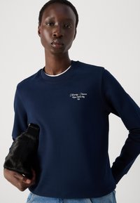 Marineblå genser med rund hals, med hvit brodert tekst "Tommy Jeans New York City" på brystet. Myk tekstur, avslappet passform.