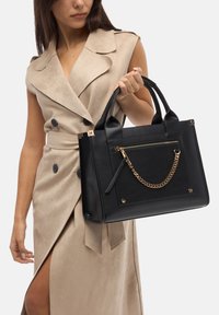 Borsa a mano in pelle nera con design strutturato, dettaglio in catena dorata e tasca frontale con zip; manici robusti. Outfit beige in suede sullo sfondo.