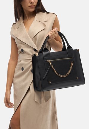 Sac en cuir noir avec un design structuré, accent en chaîne dorée et poche avant zippée ; poignées robustes. Tenue beige en suède en arrière-plan.