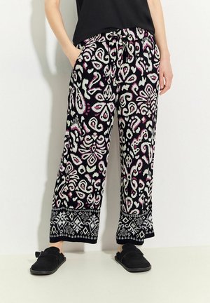 Pantalon noir à motifs avec des designs floraux blancs et roses, doté d'une taille élastique avec cordon de serrage et de poches latérales. Chaussures plates noires.
