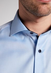 Camicia azzurra chiara con colletto aperto, bottoni scuri e tessuto testurizzato. Dettaglio ravvicinato del colletto e dell'area del collo.