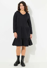 Schwarzes Hoodie-Kleid aus weichem Material mit V-Ausschnitt, langen Ärmeln und gestuftem Rüschensaum. Kombiniert mit schwarzen Reißverschluss-Stiefeletten.