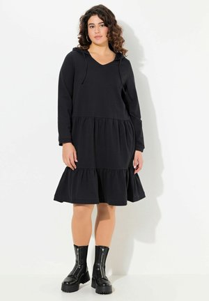 Schwarzes Hoodie-Kleid aus weichem Material mit V-Ausschnitt, langen Ärmeln und gestuftem Rüschensaum. Kombiniert mit schwarzen Reißverschluss-Stiefeletten.