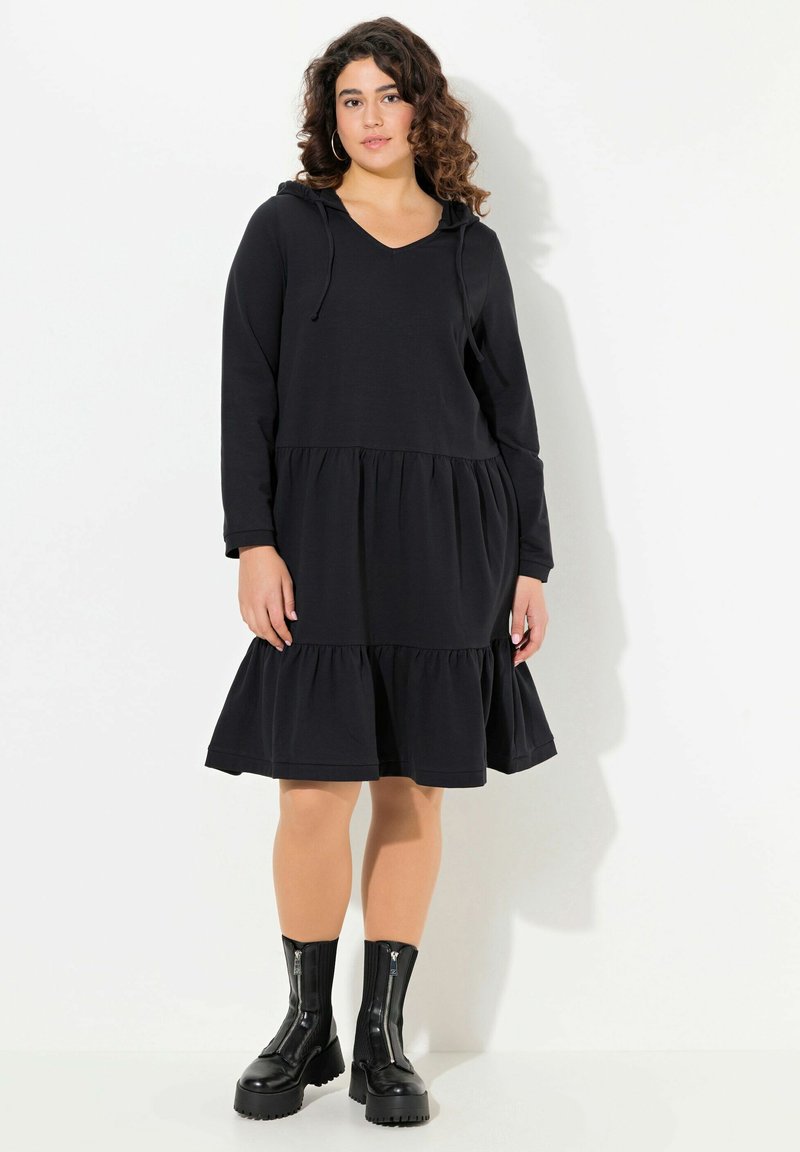 Schwarzes Hoodie-Kleid aus weichem Material mit V-Ausschnitt, langen Ärmeln und gestuftem Rüschensaum. Kombiniert mit schwarzen Reißverschluss-Stiefeletten.
