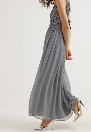 Žena v plynule se pohybujících šedých šatech s korálkovým živůtkem, obutá ve stříbrné lodičky na vysokém podpatku, v pohybu na jednobarevném pozadí.