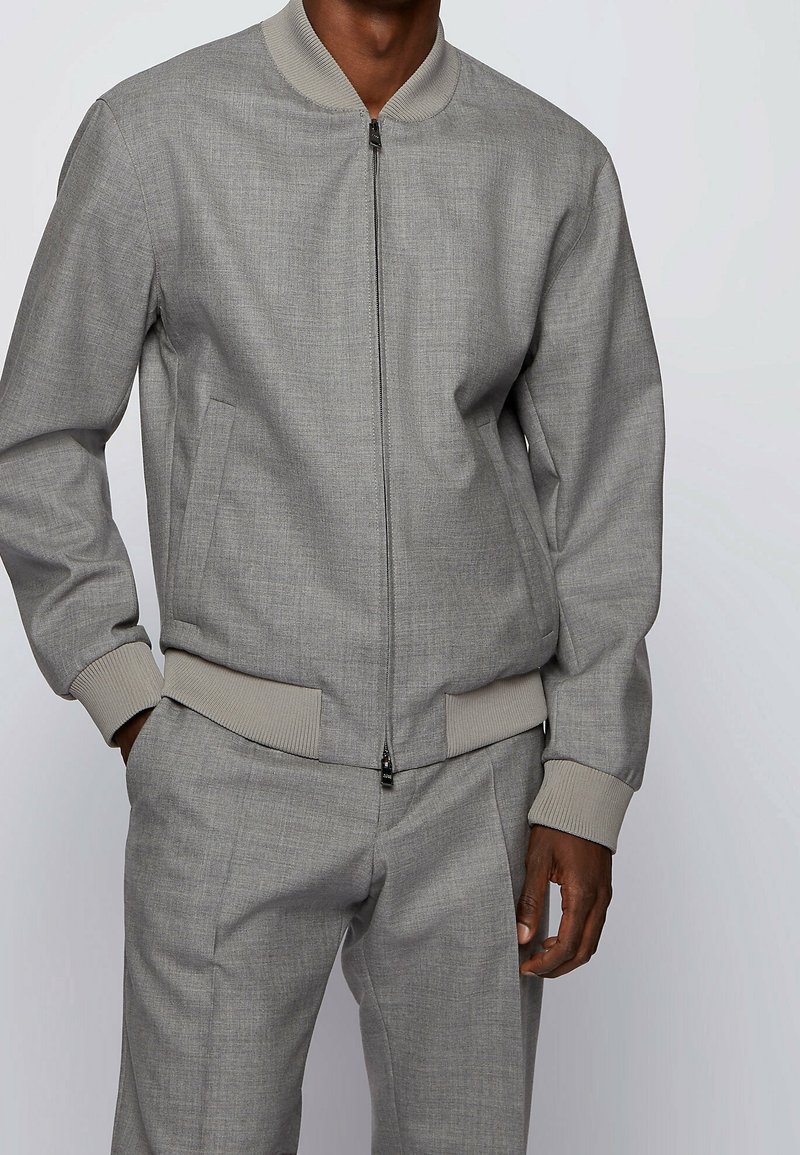Blouson aviateur gris avec finition texturée, poignets et col côtelés, et poches latérales, associé à un pantalon gris assorti.
