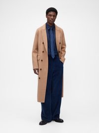 Long manteau camel à boutons croisés, porté sur une chemise à boutons bleu marine avec une cravate assortie, associé à un pantalon large en velours côtelé bleu marine.