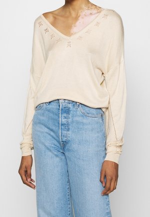 Personne portant un pull en maille crème avec de petits motifs découpés et un jean taille haute bleu clair sur un fond uni.