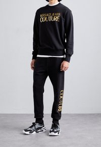 Schwarzes Sweatshirt- und Hose-Set mit goldfarbenem Schriftzug "VERSACE JEANS COUTURE". Das Material wirkt weich; die Hose hat zulaufende Bündchen. Kombiniert mit schwarzen Sneakern.