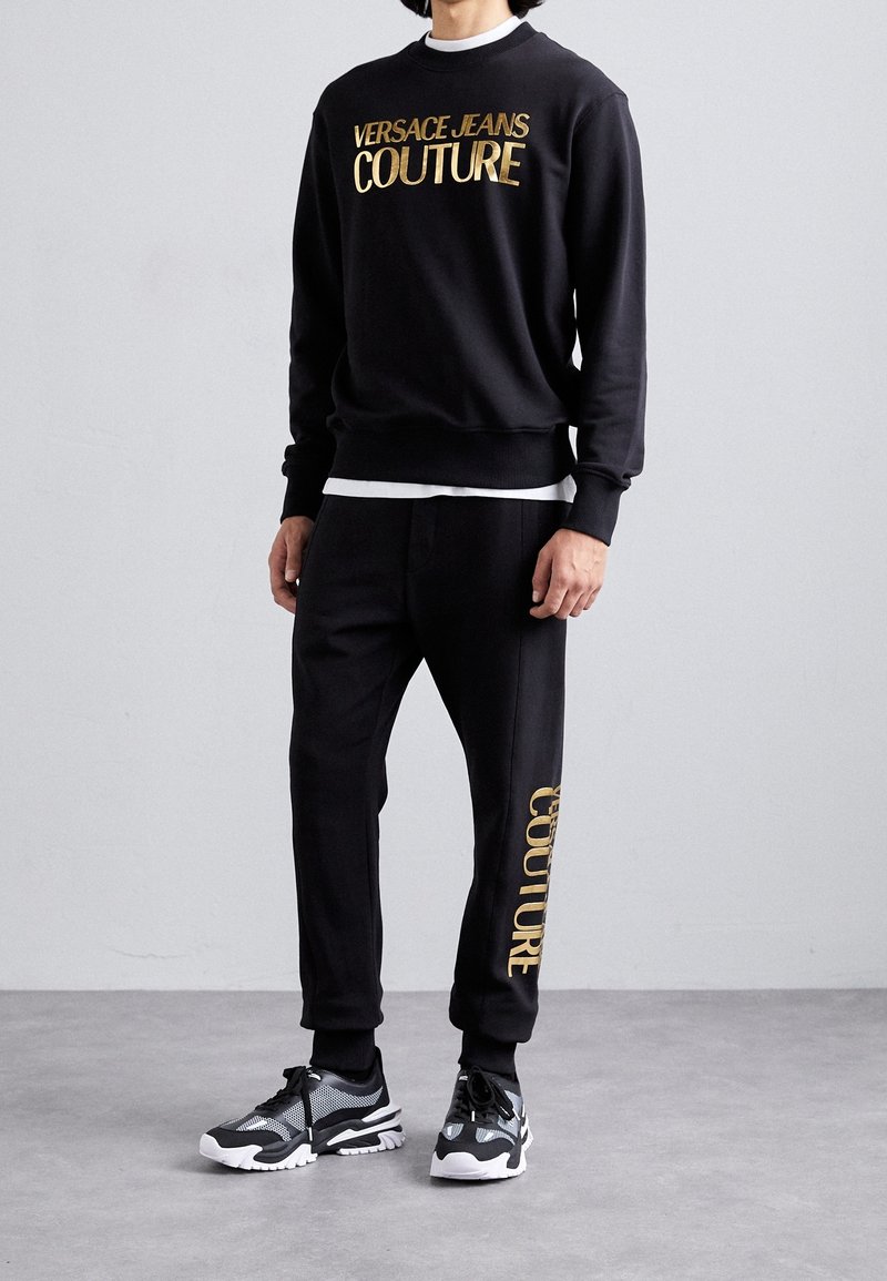 Schwarzes Sweatshirt- und Hose-Set mit goldfarbenem Schriftzug "VERSACE JEANS COUTURE". Das Material wirkt weich; die Hose hat zulaufende Bündchen. Kombiniert mit schwarzen Sneakern.