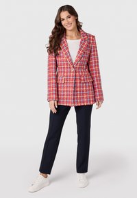 Rood en blauw geruit blazer met gesneden revers, gouden knoop en twee voorzakken, gecombineerd met donkerblauwe trousers en witte sneakers.