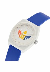 Bela ura z modrim silikonskim trakom. Okrogla številčnica z pisanimi simboli v rdeči, oranžni, rumeni in modri barvi. Številčne oznake ur in srebrna krona.