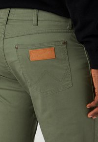 Gröna bomullsbukser med en mjuk yta, dekorerade med en tanfärgad läderlapp med "Wrangler"-logotyp och en karakteristisk zigzag-söm på bakfickan.