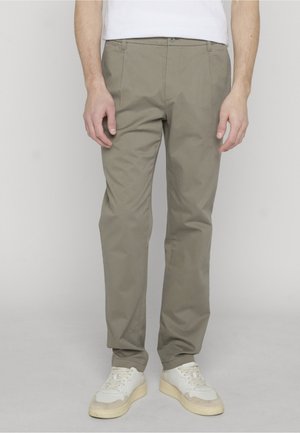 Homme portant un pantalon décontracté marron clair et des baskets blanches, debout devant un fond clair uni.